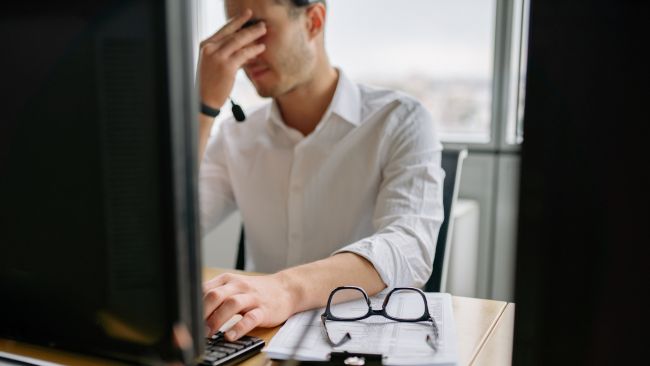 Osoba sjedi za radnim stolom u firmi ispred kompjutera i drži se za glavu jer dugotrajno sjedenje uzrokuje niz zdravstvenih problema