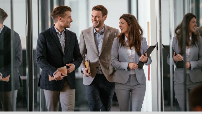 Three happy and energetic employees walking and talking together in an office hallway.
