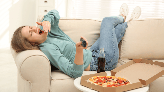 A fast-paced lifestyle affects employee health: a tired woman lying on a sofa, yawning, with a pizza and a soft drink in front of her.